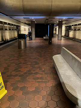 A picture of a train platform. To the left there is the bottom of a yellow wet floor cone and to the right there is a stone bench for sitting. In the background there are digital displays and pillars, as well as train tracks on the extreme left and right sides in the background. There is a ceiling at the top of the picture. A picture of a train platform. To the left there is the bottom of a yellow wet floor cone and to the right there is a stone bench for sitting. In the background there are digital displays and pillars, as well as train tracks on the extreme left and right sides in the background. There is a ceiling at the top of the picture.